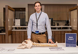 Hombre parado junto a una mesa en establecimiento médico con un modelo de columna y pelvis