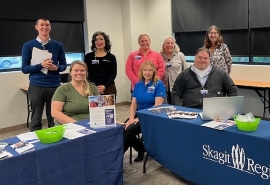 Grupo de ocho personas reunido al rededor de dos mesas con manteles de Skagit Regional Health y material promocional sobre las mesas.